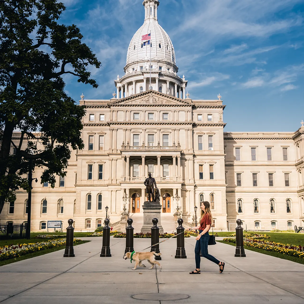 Person walking a dog in front of the State of Michigan capitol building on a sunny day.
